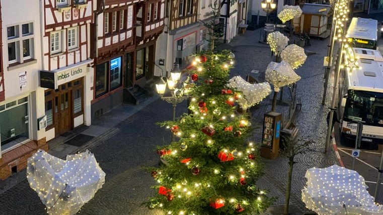 Ein festlich geschmückter Weihnachtsbaum steht auf einem gepflasterten Platz. Die Straße ist mit Lichtern und dekorativen Elementen geschmückt.