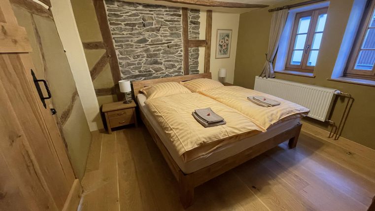 Ein gemütliches Schlafzimmer mit einem Doppelbett und Holzfußboden. Die Wände sind teilweise mit Stein und Holz gestaltet, und es gibt große Fenster, die für Tageslicht sorgen.