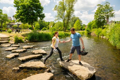 Ein Paar springt über Steine in einem klaren Bach. Im Hintergrund sind Bäume und eine grüne Landschaft zu sehen.