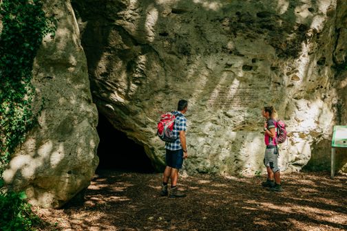 Two people are standing in front of the entrance to a cave in a wooded area.