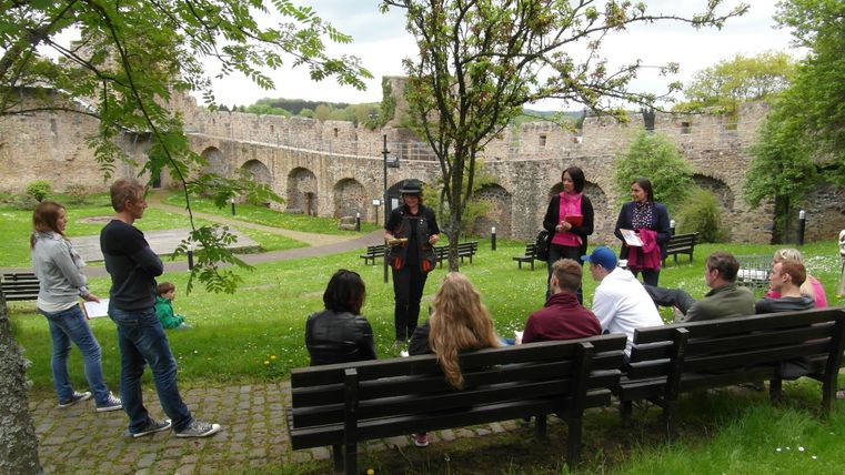 A group of hikers with their crime guide, some sitting on benches, some standing in front of the backdrop of the city wall in Hillesheim.