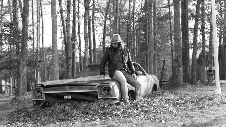 A man is sitting on an old, rusty car that is covered with leaves. In the background, there are trees that create an autumn atmosphere.