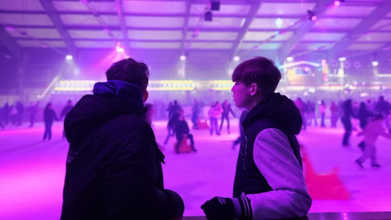 Twee personen staan aan de rand van een kunstijsbaan en observeren de schaatsers. Kleurrijke lichten zorgen voor een feestelijke sfeer.