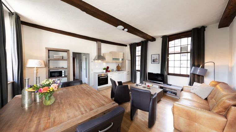 Ein stilvolles Wohnzimmer mit einem großen Holzspeisetisch und bequemen schwarzem Mobiliar. Die moderne Küche ist offen gestaltet und das Licht strömt durch große Fenster.