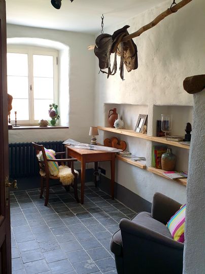 A cozy room with a desk, a comfortable armchair, and shelves. Some hats hang on the wall, and a window provides natural light.