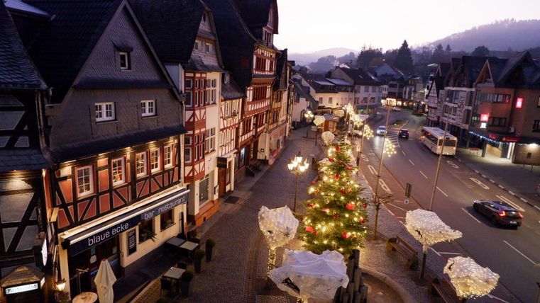 Eine festlich geschmückte Straße mit einem beleuchteten Weihnachtsbaum. Historische Gebäude und sanfte Hügel im Hintergrund schaffen eine gemütliche Atmosphäre.