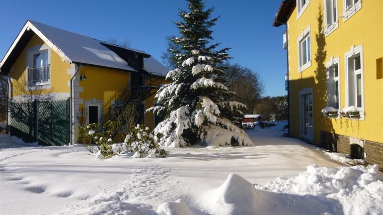 Ein verschneiter Hof mit einem großen Tannenbaum und gelben Häusern im Hintergrund. Der Himmel ist klar und blau, was eine gemütliche Winteratmosphäre schafft.