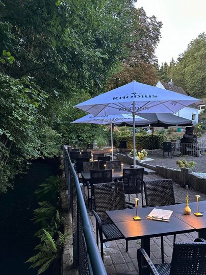 A cozy restaurant by the river with dark tables and chairs. Umbrellas provide shade from the sun, and the surroundings are green and inviting.