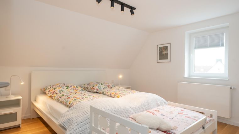 Ein helles Schlafzimmer mit einem großen Bett und einem zusätzlichen Kinderbett. Die Wände sind hell, und ein Fenster lässt viel Tageslicht herein.