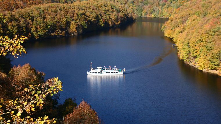 A boat sails on a calm lake, surrounded by colorful trees in autumn. The landscape is picturesque and reflects the colors of nature.