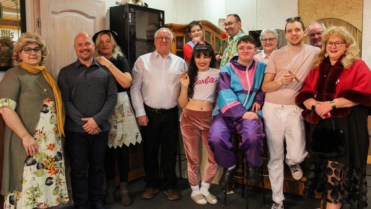 A group of people is standing together in a room. They are wearing colorful and diverse clothing and seem to be having fun.