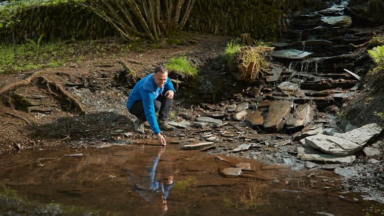 Ein Mann in blauer Jacke hockt an einem kleinen Bachlauf im Wald und berührt das Wasser mit der Hand; im Hintergrund fließt Wasser über flache Schieferplatten.