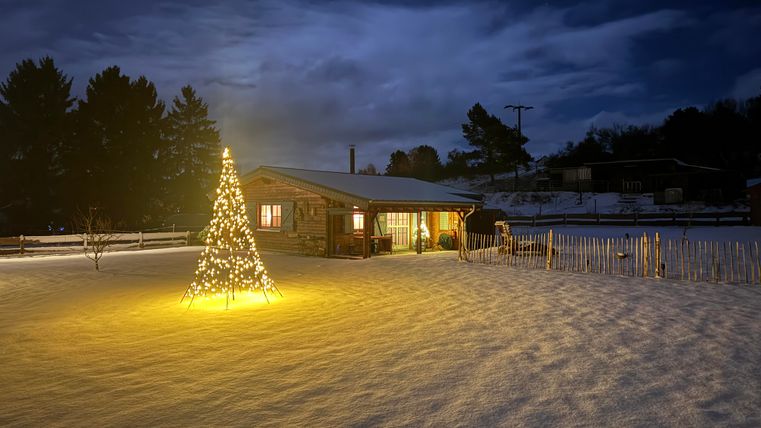 A cozy house in the snow at night, surrounded by a sparkling light tree. The sky is overcast and the atmosphere is calm and festive.