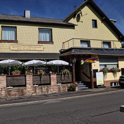 Ein gemütliches Gasthaus mit Außenbereich und Sonnenschirmen. Im Hintergrund ist eine ruhige Straße und ein blauer Himmel zu sehen.