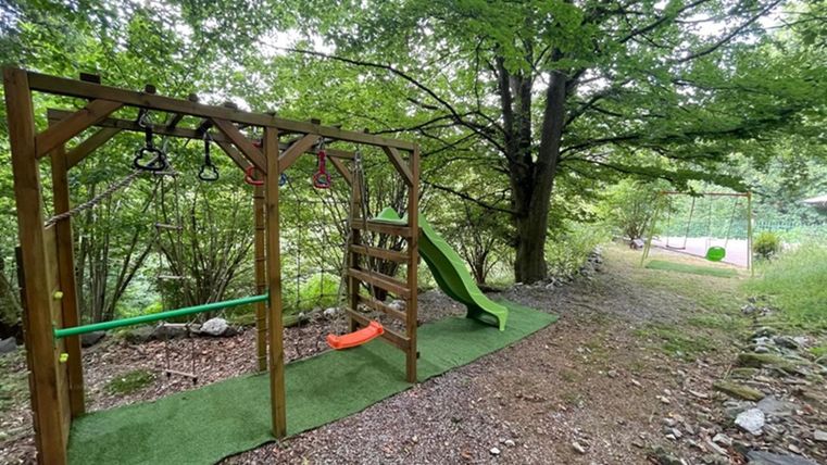 A playground structure made of wood with a slide and climbing elements is surrounded by trees. The ground is covered with green carpet and there is a narrow path in the background.