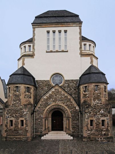 Een moderne kerk met een markante, ronde toren en stenen rondbogen. De ingang is ruim opgezet met trappen en de gevel is in lichte kleuren uitgevoerd.