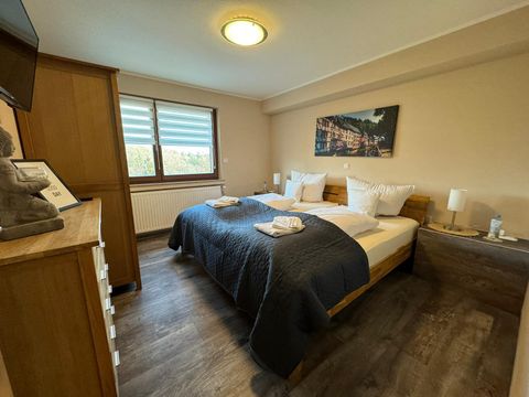 A cozy bedroom with a large bed and modern furniture. The walls are in soft colors and large windows let in plenty of light.