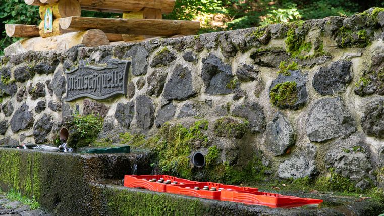 A wall on which hangs a sign with the inscription "Maischquelle". On the wall is a wooden table with two benches. Empty glass bottles and crates of drinks lie under the wall.