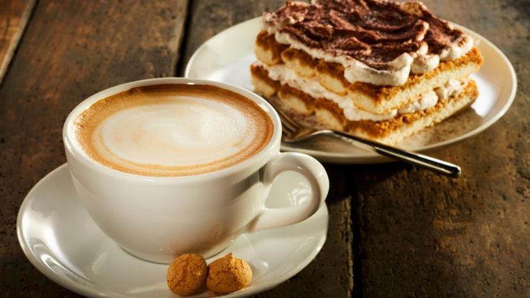 Eine Tasse Cappuccino mit zwei Keksen auf einer Untertasse und ein Stück Tiramisu auf einem Teller.