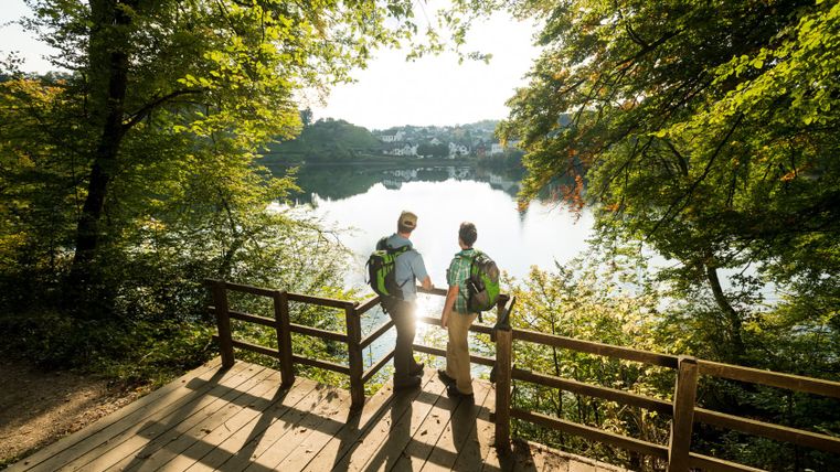 Twee mensen staan op een houten loopbrug en kijken uit over een meer omringd door bomen.