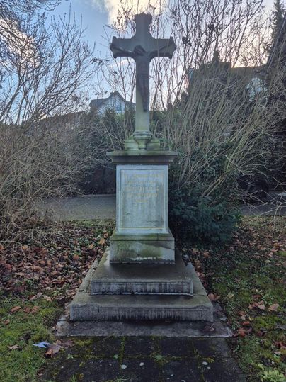 Une vieille pierre tombale avec une croix se dresse dans une zone verdoyante. Entourée de branches sèches et d'herbe, la scène est calme et contemplative.
