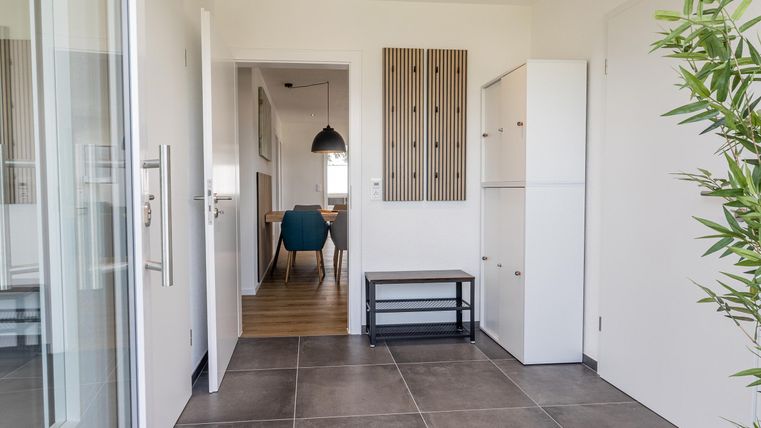 Een moderne entree met grijze tegels en een lichte wandafwerking. Op de achtergrond zijn een tafel en stoelen zichtbaar.