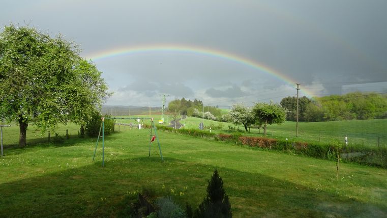 Een groene tuin met een regenboog die over het landschap spanning. Op de achtergrond zijn bomen en weilanden te zien.