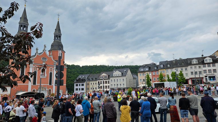 Ein belebter Platz mit einer Kirche im Hintergrund und vielen Menschen. Der Himmel ist bewölkt und die Atmosphäre wirkt festlich.