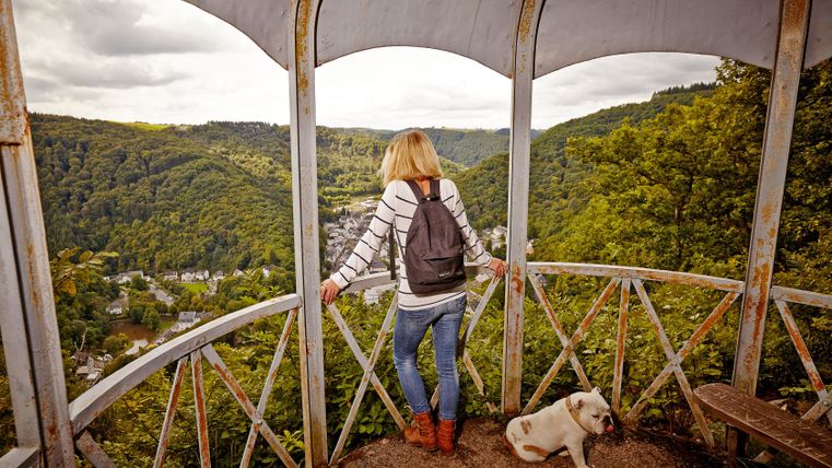 Frau mit Hund auf einem Aussichtsturm, Blick auf bewaldetes Tal und Dorf.