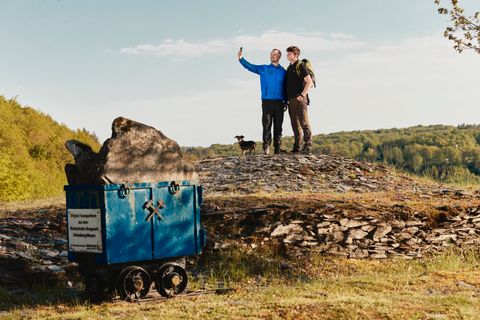 Twee wandelaars met een hond nemen een selfie op een leisteenveld in de Eifel. Links op de voorgrond staat een blauwe transportwagen met leisteen, daarachter bossen en groene heuvels onder een blauwe lucht.