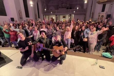 A lively music event with a large crowd of enthusiastic people. The artists on stage play instruments and the audience celebrates with raised hands.