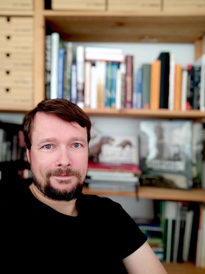 Ein Mann mit Bart sitzt vor einem Bücherregal. Im Hintergrund sind zahlreiche Bücher und einige Boxen zu sehen.