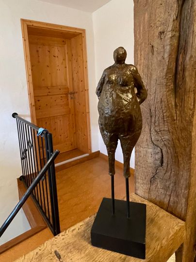 Eine sculpturale Figur aus Bronze steht auf einem Sockel. Im Hintergrund ist eine Holztür und eine Treppe zu sehen.