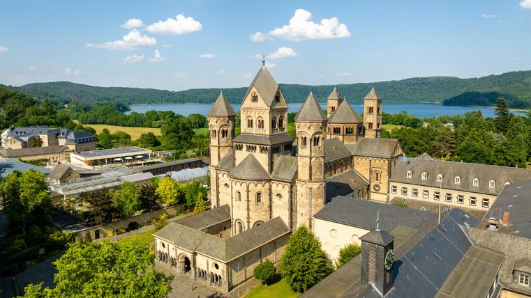 Een indrukwekkend kloostercomplex met markante torens, omringd door weelderig groen. Op de achtergrond zijn het meer en het heuvelachtige landschap zichtbaar.