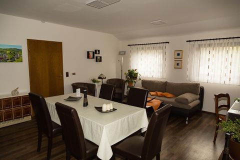 A cozy living area with a dining table and chairs. Light pours in through the windows, and there is a sofa corner with cushions.