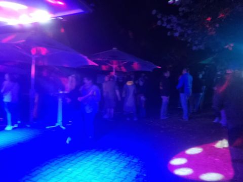 Eine bunte Party mit Musik und Lichtern im Freien. Viele Menschen stehen unter Schirmen und genießen die Atmosphäre.