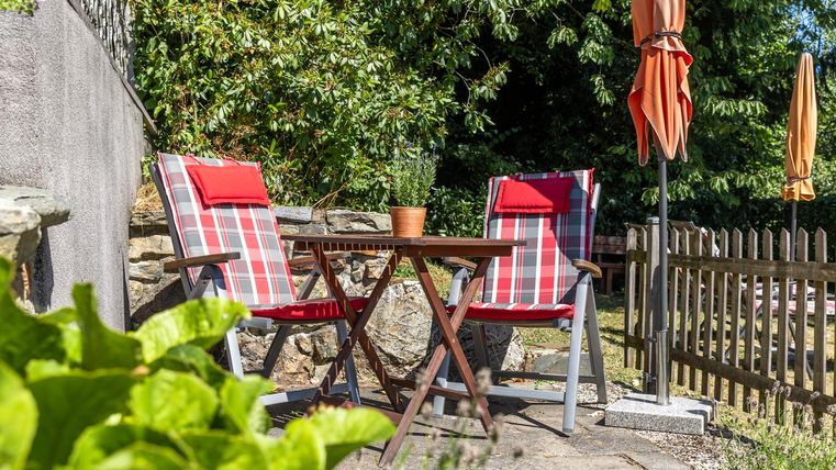 An inviting garden area with two striped chairs and a small table. In the background, there are umbrellas and a wooden slatted fence frames the space.