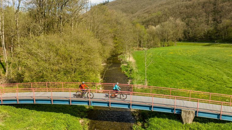 Deux cyclistes sur un pont enjambant une rivière, entourés de prairies verdoyantes et de collines boisées.