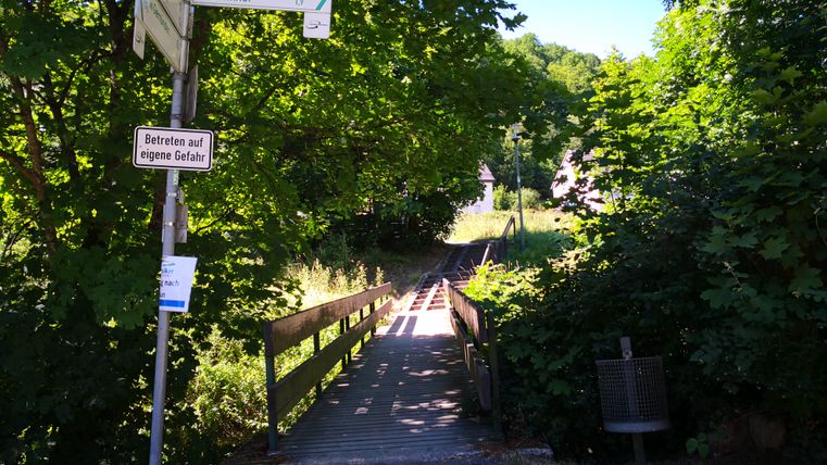 A narrow wooden walkway leads through a green, wooded area. Signs point to the center and the train station.
