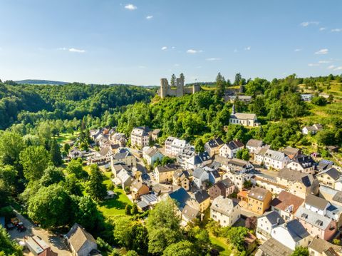 A picturesque view of a small village in a wooded valley. In the background, an old castle towers over the charming architecture of the houses.