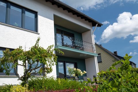 A modern house with a balcony and large windows. Surrounded by green plants and a blue sky.