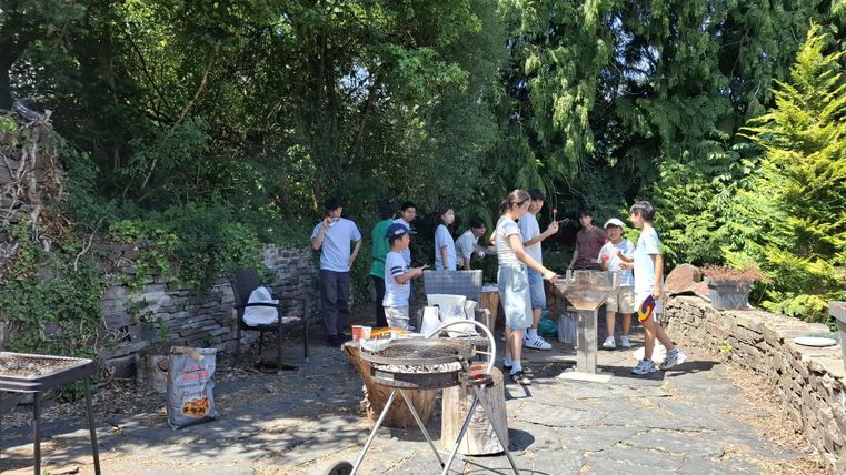 Eine Gruppe von Menschen bereitet ein Grillfest im Freien vor. Es gibt einen Grill und einige Tische auf einer steinernen Fläche, umgeben von üppigem Grün.