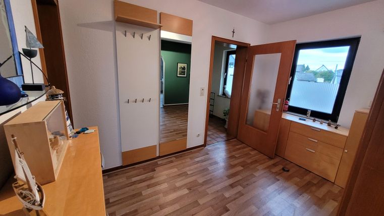 Ein heller Flur mit Holzfußboden und modernen Möbeln. Im Hintergrund sind grüne Wände und ein Fenster sichtbar.