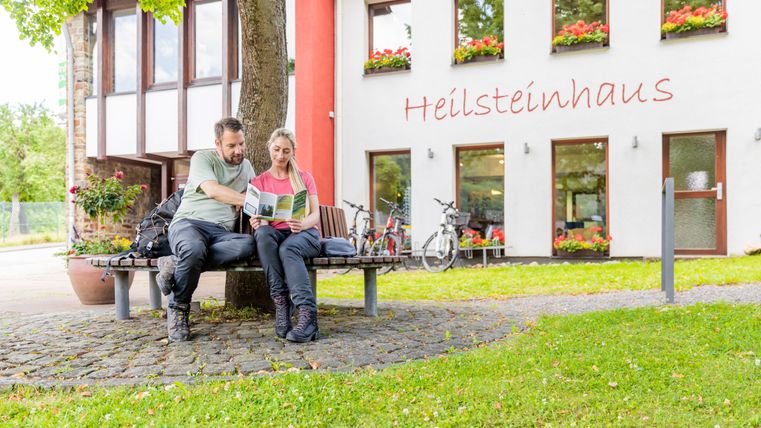 Two people sit on a bench in front of the Heilsteinhaus in Einruhr and read a brochure.