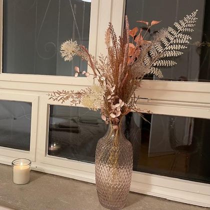 Eine elegante Vase mit getrockneten Blumen steht auf einem Fensterbrett. Neben der Vase befindet sich eine brennende Kerze.