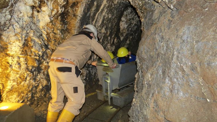 A miner pushes a cart in a mine shaft. The walls are made of rock and the lighting is dim.