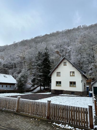 A white house stands at the foot of a snow-covered slope. The surroundings are quiet and wintry, with snow-covered trees in the background.