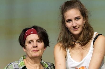 Zwei Frauen stehen auf einer Bühne. Die eine trägt ein kariertes Hemd und ein rotes Stirnband, die andere hat lange Haare und trägt ein weißes Top.