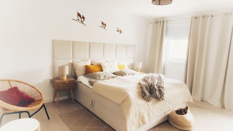 Une chambre lumineuse avec un grand lit et des coussins décoratifs. Il y a une chaise confortable et beaucoup de lumière naturelle par la fenêtre.