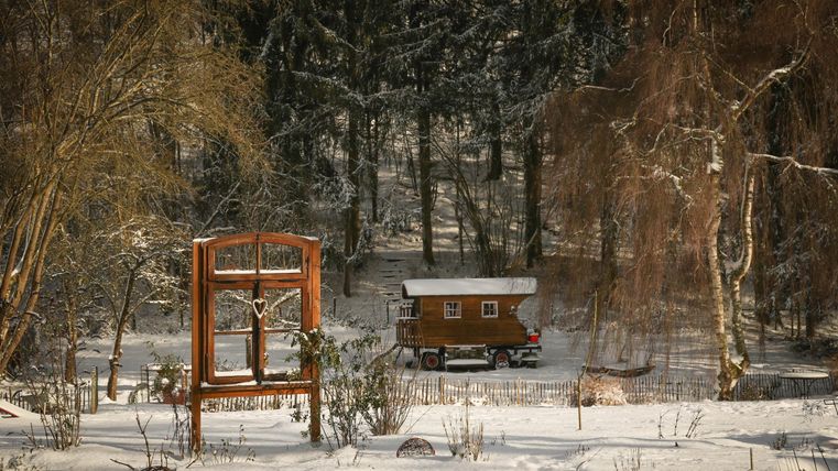 Ein hübsches Holzhaus steht in einer verschneiten Landschaft umgeben von Bäumen. Im Vordergrund ist ein rustikaler Holzzaun mit einer Fensterinstallation zu sehen.